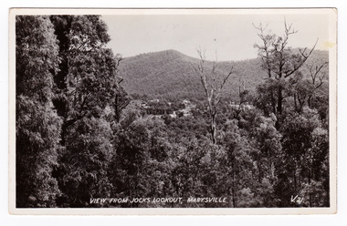 Shows the view from Jock's Lookout of Marysville in Victoria. In the distance can be seen various buildings in Marysville. In the foreground are several trees and in the background are heavily forested mountains. The title of the photograph is handwritten in white ink on the lower edge. On the reverse of the postcard is space to write a message and an address and to place a postage stamp. The postcard is unused.