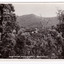 Shows the view from Jock's Lookout of Marysville in Victoria. In the distance can be seen various buildings in Marysville. In the foreground are several trees and in the background are heavily forested mountains. The title of the photograph is handwritten in white ink on the lower edge. On the reverse of the postcard is space to write a message and an address and to place a postage stamp. The postcard is unused.