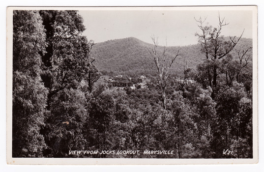 Shows the view from Jock's Lookout of Marysville in Victoria. In the distance can be seen various buildings in Marysville. In the foreground are several trees and in the background are heavily forested mountains. The title of the photograph is handwritten in white ink on the lower edge. On the reverse of the postcard is space to write a message and an address and to place a postage stamp. The postcard is unused.
