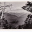Shows the view of Marysville taken from Mount Gordon. In the distance can be seen the buildings of Marysville surrounded by heavily forested mountains. In the foreground are some large trees. The title of the photograph is handwritten in white in on the lower edge. On the reverse of the postcard is a space to write a message and an address and to place a postage stamp. The postcard is unused.