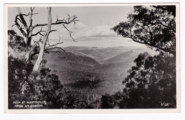 Shows the view of Marysville taken from Mount Gordon. In the distance can be seen the buildings of Marysville surrounded by heavily forested mountains. In the foreground are some large trees. The title of the photograph is handwritten in white in on the lower edge. On the reverse of the postcard is a space to write a message and an address and to place a postage stamp. The postcard is unused.