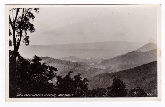 Shows the view of Marysville in Victoria taken from Nicholl's Lookout. In the distance can be seen the buildings of Marysville surrounded by heavily forested mountains. The title of the photograph is handwritten in white ink on the lower edge. On the reverse of the postcard is a space to write a message and an address and to place a postage stamp. The postcard is unused.