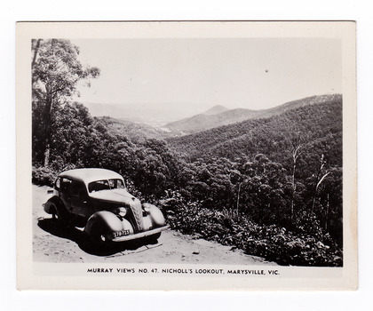 Shows the view of Marysville taken from Nicholl's Lookout. In the foreground is shown an early model car. Shown in the background are a series of heavily forested mountains. The title of the photograph is shown along the lower edge of the photograph.
