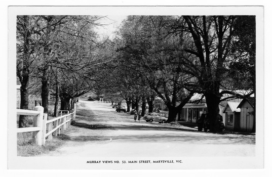 Shows the main street in Marysville, Victoria. Shows an unsealed road with buildings along the right hand side. There are several cars parked on the edge of the road facing the buildings. Behind one of the cars can be seen two ladies. The left hand side of the road is lined by wooden fences. On both sides of the road can be seen several large trees. The title of the postcard is shown along the lower edge of the postcard. On the reverse of the postcard is a space to write a message and an address and to place a postage stamp. The postcard is unused.
