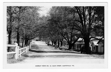 Shows the main street in Marysville, Victoria. Shows an unsealed road with buildings along the right hand side. There are several cars parked on the edge of the road facing the buildings. Behind one of the cars can be seen two ladies. The left hand side of the road is lined by wooden fences. On both sides of the road can be seen several large trees. The title of the postcard is shown along the lower edge of the postcard. On the reverse of the postcard is a space to write a message and an address and to place a postage stamp. The postcard is unused.