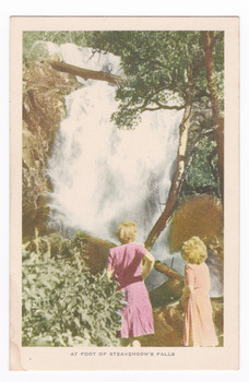 Shows two ladies standing at the foot of Steavenson Falls in Maryville in Victoria. Shows the ladies looking at the bottom tier of the waterfalls cascading large boulders. The waterfalls are surrounded by forest and across the top of the tier there is a fallen log. The title of the postcard is shown along the lower edge of the postcard. On the reverse of the postcard is a space to write a message and an address and to place a postage stamp. The postcard is unused.