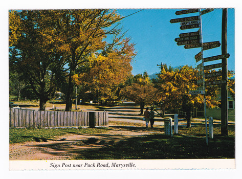 Shows the sign post that stood opposite Pack Road in Marysville in Victoria. Shows the signpost in the foreground. Opposite the signpost is a wooden picket fence. There are two people walking past the signpost. The title of the postcard is shown along the lower edge of the postcard. On the reverse of the postcard is a space to write a message and an address and to place a postage stamp. The postcard is unused.