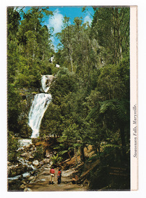 Shows Steavenson Falls in Marysville in Victoria. Shows the falls cascading down the mountain. The falls are surrounded by forest. Shows the track leading to the falls. On the track there are two people standing looking at the falls. On the reverse of the postcard is a space to write a message and an address and to place a postage stamp. The postcard is unused.