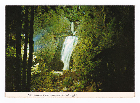 Shows Steavenson Falls in Marysville in Victoria, illuminated at night. Shows the falls cascading down the mountain which is heavily forested. On the reverse of the postcard is a space to write a message and an address and to place a postage stamp. The postcard is unused.