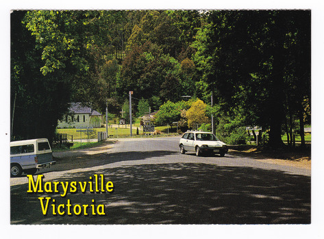 Shows the tree lined main street in Marysville in Victoria. The road leads to a roundabout. In the background can be seen a church with the rectory beside it. On the reverse of the postcard is a space to write a message and an address and to place a postage stamp. The postcard is unused.