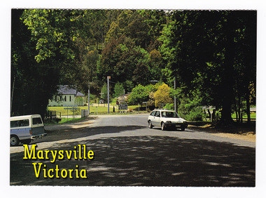 Shows the tree lined main street in Marysville in Victoria. The road leads to a roundabout. In the background can be seen a church with the rectory beside it. On the reverse of the postcard is a space to write a message and an address and to place a postage stamp. The postcard is unused.