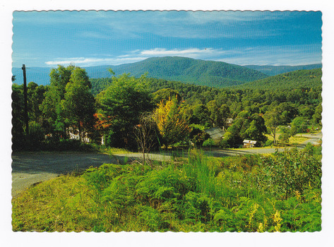Shows the view of Marysville in Victoria from Sheehan's Lookout. In the foreground is a road flanked by some houses. In the background are a series of heavily forested mountains. On the reverse of the postcard is a space to write a message and an address and to place a postage stamp. The postcard is unused.