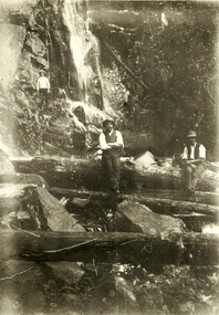 Shows a group of three men sitting and standing at the base of Steavenson Falls in Marysville in Victoria. The men are sitting on rocks and standing on large logs that are across the base of the falls.