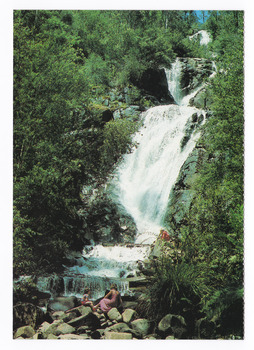 Shows Steavenson Falls in Marysville in Victoria. Shows two adults and one child sitting on rocks at the base of the falls. On the reverse of the postcard is a space to write a message and an address and to place a postage stamp. The postcard is unused.