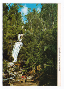 A colour photograph of Steavenson Falls in Marysville in Victoria. Photograph shows two people standing on a track at the base of the falls. On the reverse of the postcard is a space to write a message and an address and to place a postage stamp. The postcard is unused.