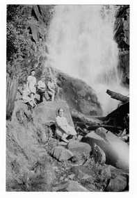 Shows a group of people sitting at the base of Steavenson Falls in Marysville in Victoria. On the reverse of the photograph is a sticker detailing the location and date the photograph was taken.