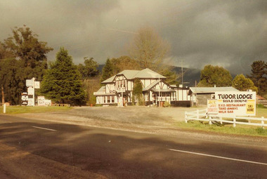 Shows Tudor Lodge Roadhouse in Narbethong in Victoria. Shows a large tudor style building with a petrol bowser situated out the front of the building. In the right of the photograph is a large sign advertising the roadhouse, restaurant, take away, milk bar and ski hire. On the roadside at the front of the building is another sign advertising what is available at the roadhouse.