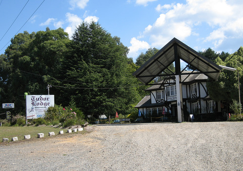 Shows Tudor Lodge Roadhouse in Narbethong in Victoria. Shows a large tudor style building with a petrol bowser situated out the front of the building. On the roadside at the front of the building is another sign advertising what is available at the roadhouse including take away food and ski hire.