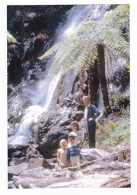 Shows a man and three children standing at the base of Steavenson Falls in Marysville in Victoria. Behind the group are two tree ferns.