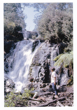 Shows two ladies, one sitting and one standing, at the base of Steavenson Falls in Marysville in Victoria. Shows the falls cascading down the mountain surrounded by forest.