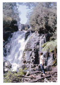 Shows two ladies, one sitting and one standing, at the base of Steavenson Falls in Marysville in Victoria. Shows the falls cascading down the mountain surrounded by forest.