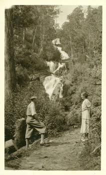 Shows a man and a women standing on the track that leads to the base of Steavenson Falls in Marysville in Victoria. The falls can be seen in the background.