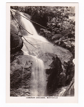 Shows the Cameron Cascades near Marysville in Victoria. Shows the waterfall cascading down over some large boulders. In the background can be seen trees and tree ferns. The title of the photograph is along the lower edge of the photograph.