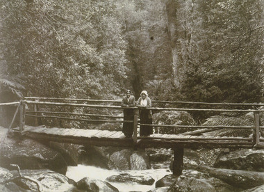Shows two ladies standing on a timber bridge crossing the Taggerty River. The river is flowing over large boulders and is surrounded by a forest of large trees and tree ferns.