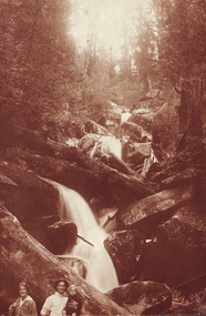 Shows a woman and a man who is holding a small child standing at the base of Keppel Falls near Marysville in Victoria. In the background the falls are cascading over large boulders and lying across the falls are several large logs.