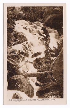 Shows the Cora Lynn Falls near Marysville in Victoria. Shows the falls cascading down the mountain surrounded by several boulders. There is a fallen log across the falls. On the reverse of the postcard is a space to write a message and an address and to place a postage stamp. The postcard is unused.