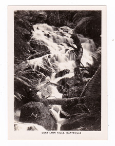Shows the Cora Lynn Falls near Marysville in Victoria. Shows the falls cascading down the mountain surrounded by several boulders. There is a fallen log across the falls. The title of the photograph is along the lower edge of the photograph.