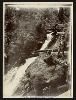 Shows two men, one sitting and one standing, at the edge of the waterfall which is cascading down the mountain. There are a few fallen logs lying across the falls and the falls are surrounded by forest.
