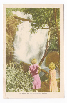 Shows two ladies standing at the foot of Steavenson Falls in Maryville in Victoria. Shows the ladies looking at the bottom tier of the waterfalls cascading large boulders. The waterfalls are surrounded by forest and across the top of the tier there is a fallen log. The title of the postcard is shown along the lower edge of the postcard. On the reverse of the postcard is a space to write a message and an address and to place a postage stamp. The postcard is unused.