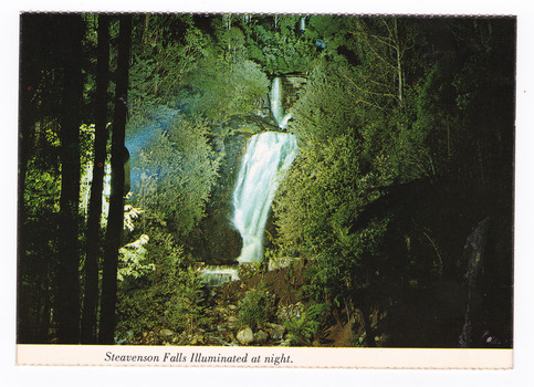 Shows Steavenson Falls in Marysville in Victoria, illuminated at night. Shows the falls cascading down the mountain surrounded by forest. On the reverse of the postcard is a space to write a message and an address and to place a postage stamp. The postcard is unused.
