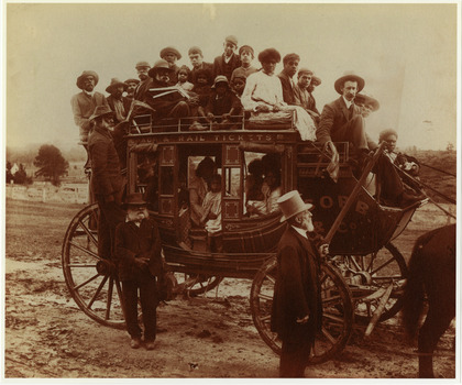 Shows a large group of Aboriginal men, women and children on board a Cobb & Co Coach. The coach driver is a white man. Two of the Aboriginal men are carrying boomerangs. All the aboriginal people are wearing western dress; the men are in suits and hats. 