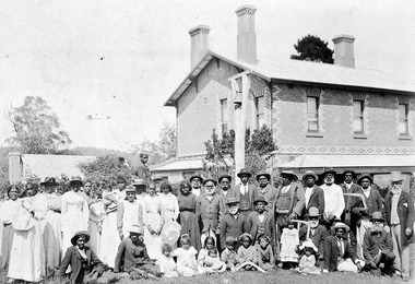 Shows a large group of Aboriginal men, women and children standing and sitting in front of a two-storey brick building. Most of the men are wearing suits and hats and the women are all wearing long dresses, some with hats. The children are wearing similar outfits.
