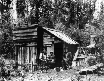 Shows three men sitting outside a wooden hut in the forest. With the men is a dog. The men are eating. Leaning against the hut is an axe. In the background can be seen another wooden building.