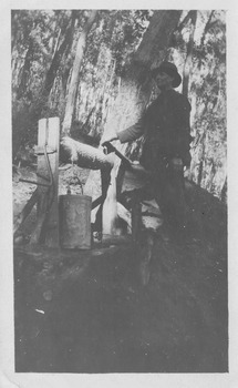Shows a man standing alongside a well turning the handle to raise the bucket. He is surrounded by forest.