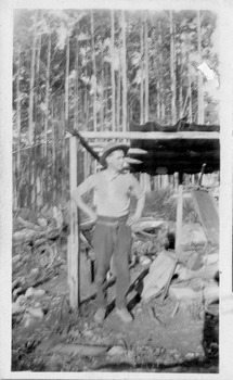 Shows a man standing next to the blacksmith's forge at the Golden Bower mine in the Cumberland Valley in Victoria. The forge consists of four poles with some cross poles and a corregated iron roof. The forge is surrounded by forest.