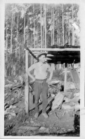 Shows a man standing next to the blacksmith's forge at the Golden Bower mine in the Cumberland Valley in Victoria. The forge consists of four poles with some cross poles and a corregated iron roof. The forge is surrounded by forest.