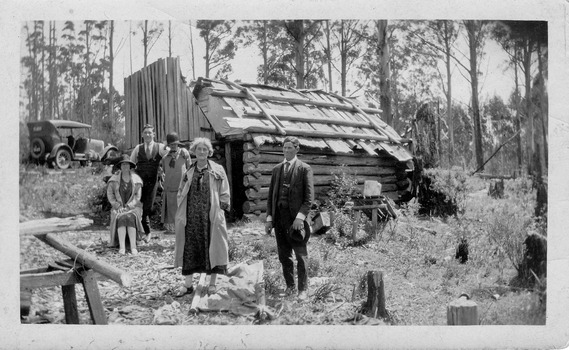 Shows a group of three women and two men standing and sitting outside a large wooden hut. In the foreground there is a wooden saw horse. In the background there is an early model car in amongst the forest.