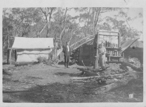 Shows a man and a woman standing near the mine shaft head of the Golden Bower mine in the Cumberland Valley. The mine head has been secured with several logs and rocks. In the background can be seen a large white tent.