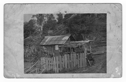 Shows a man sitting outside a wooden hut. The man is smoking. The hut is surrounded by a wooden paling fence and at the of the hut is a lean-to. Both the hut and the lean-to are roofed with timber palings. In the background is a steep hill with wooden paling fences running up the slope. On the reverse of the postcard is a hand-written message and an address.