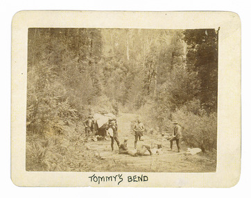 Shows a group of six men sitting and standing in the middle of a corduroy road that leads through the forest. One of the men is standing at the rear of a carriage. In the background can be seen a campfire with a billy can suspended above it. The men are all dressed in suits and hats. The road is surrounded by forest. The title of the photograph is along the lower edge in black ink. On the reverse of the photograph is a stamp from the Armstrong Collection at 42 Station Street in Sandringham in Victoria.