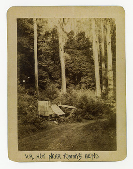Shows a small timber hut by the side of a dirt road which leads through a forest of large trees. The title of the photograph is along the lower edge in black ink. On the reverse of the photograph is a stamp from the Armstrong Collection at 42 Station Street in Sandringham in Victoria.