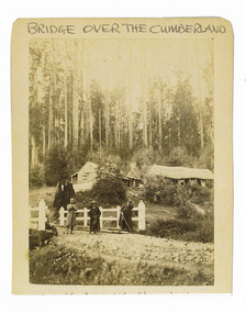 Shows a group of four men standing and sitting on a white painted wooden bridge. In the background, backing onto the forest, are two wooden buildings with a large chimney set between them. The title of the photograph is along the top edge in black ink. On the reverse of the photograph is a stamp from the Armstrong Collection at 42 Station Street in Sandringham in Victoria.