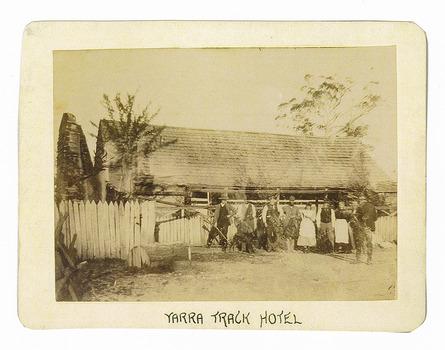 Shows a timber shingled building with a group of nine people and one dog standing outside. One of the people has his back turned to the camera. On the right hand side of the building is a large brick chimney. Running along the front of the building is a white painted picket fence. The title of the photograph is along the lower edge in black ink. On the reverse of the photograph is a stamp from the Armstrong Collection at 42 Station Street in Sandringham in Victoria.