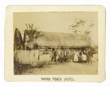 Shows a timber shingled building with a group of nine people and one dog standing outside. One of the people has his back turned to the camera. On the right hand side of the building is a large brick chimney. Running along the front of the building is a white painted picket fence. The title of the photograph is along the lower edge in black ink. On the reverse of the photograph is a stamp from the Armstrong Collection at 42 Station Street in Sandringham in Victoria.