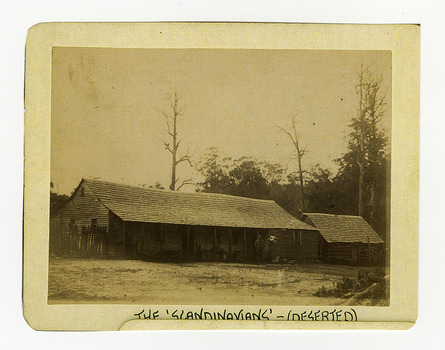 Shows the rear view of The Scandinavians' which was a hotel and boarding house along the Yarra Track. Shows a timber clad building with a timber shingled roof. At the right of the building is a wooden picket fence and at the left of the building is another timber clad building. Under the verandah of the main building can be seen various items. There is a man standing in front of the building. The title of the photograph is along the lower edge in black ink. On the reverse of the photograph is a stamp from the Armstrong Collection at 42 Station Street in Sandringham in Victoria.