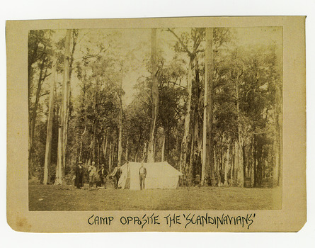 Shows tent set up in a clearing in the forest. Standing outside of the tent are five men. One of the men appears to have what appears to be an axe resting on his right shoulder. The title of the photograph is along the lower edge in black ink. On the reverse of the photograph is a stamp from the Armstrong Collection at 42 Station Street in Sandringham in Victoria.